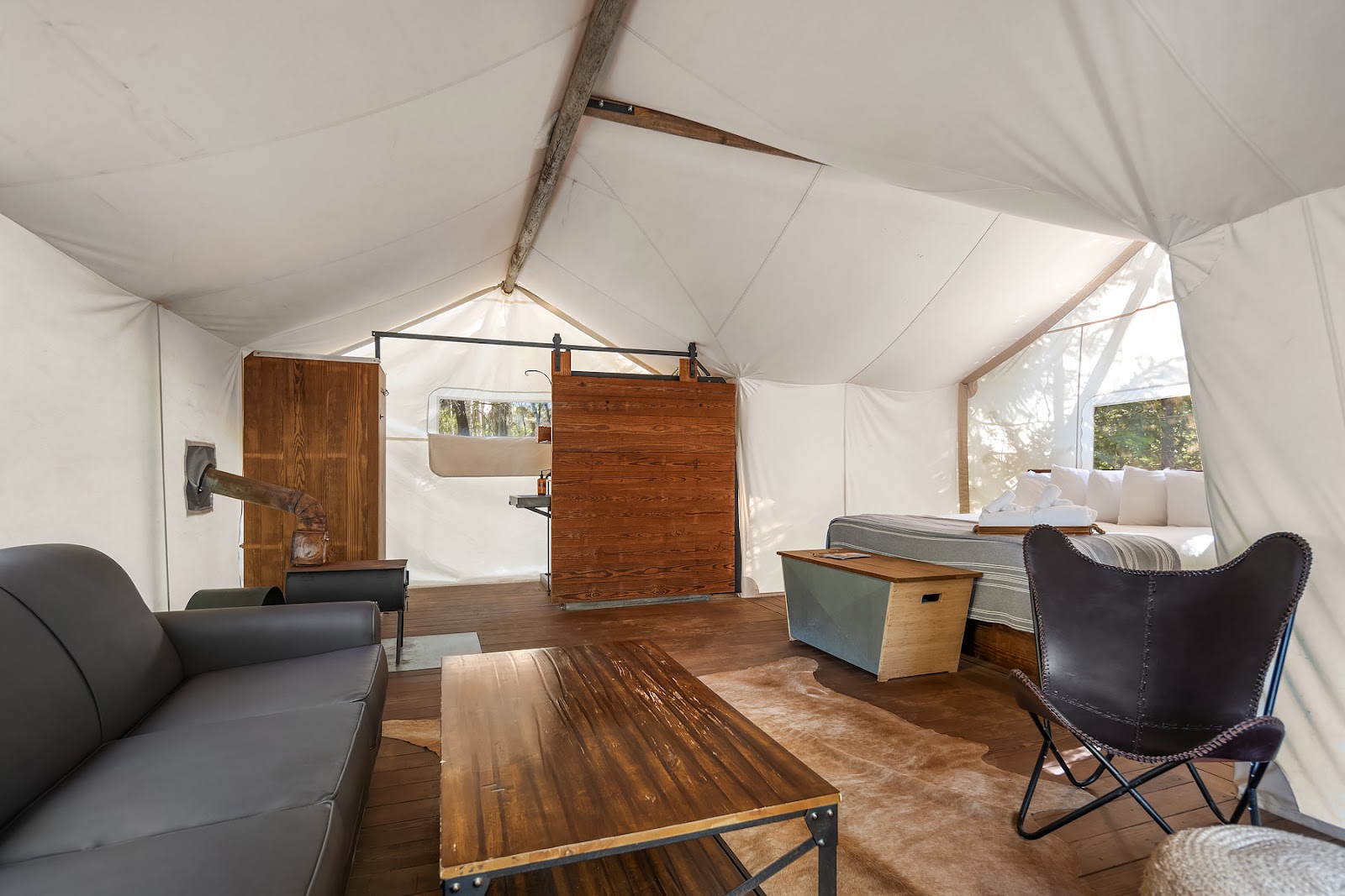 Spacious tent interior in Glacier National Park with a wooden bed, leather chair, gray sofa, coffee table, and a rustic stove pipe.