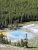 Bright blue geothermal pool with mineral rims and steam, beside a pine forest and grassy trail in Yellowstone National Park.