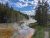 Steam rises from mineral-colored geothermal pools along a forested stream with a dirt trail and a hiker on the right, in Yellowstone National Park.