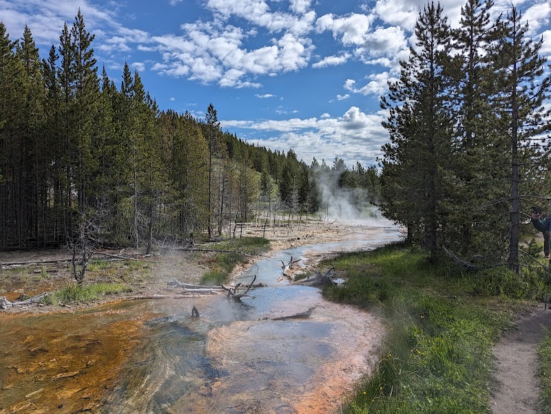 Steam rises from mineral-colored geothermal pools along a forested stream with a dirt trail and a hiker on the right, in Yellowstone National Park.