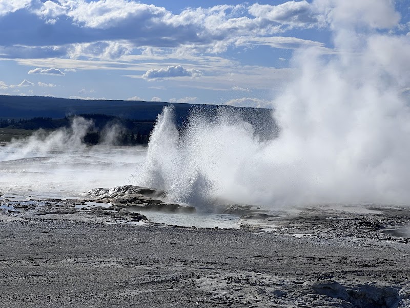 Yellowstone National Park features a geyser erupting, sending a tall steam plume over a rocky geothermal field.