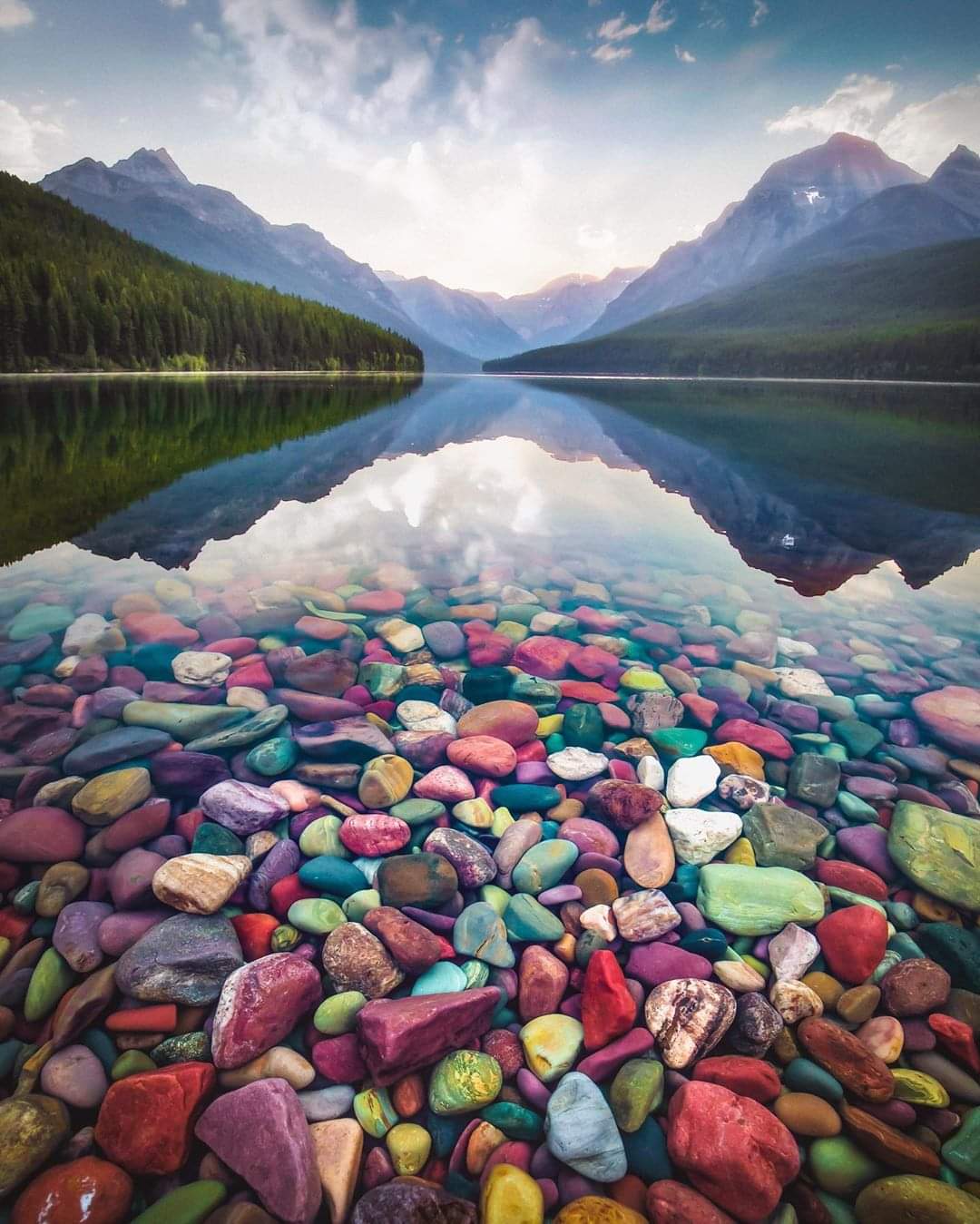 Numa Peak mirrored in a calm glacier-fed lake at Glacier National Park, with rainbow-colored pebbles in the foreground.