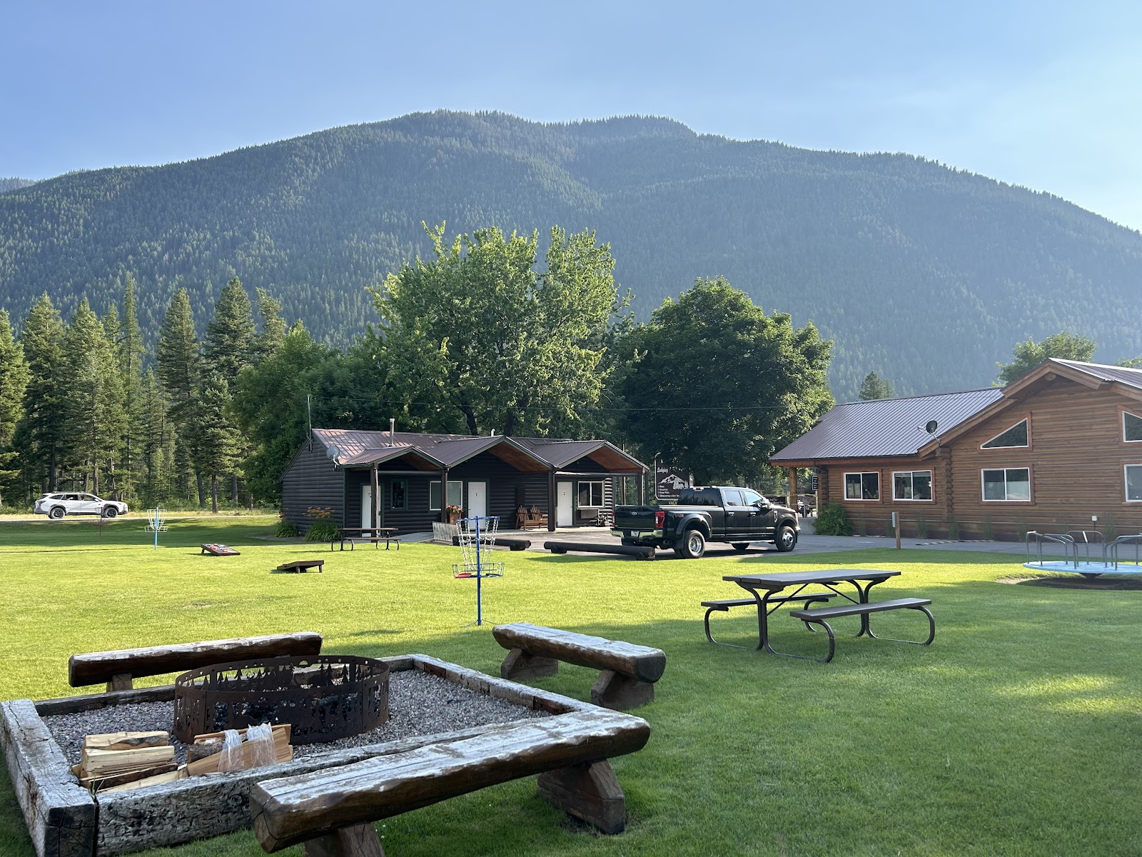 Cabin-style lodging at Beargrass Lodging and RV Resort nestled in Glacier National Park, with green lawn, trees, and forested mountains in the background.