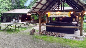 Rustic open-air cafe under a wooden pavilion at Glacier National Park's campground, with picnic tables and a wood-fired grill nearby.