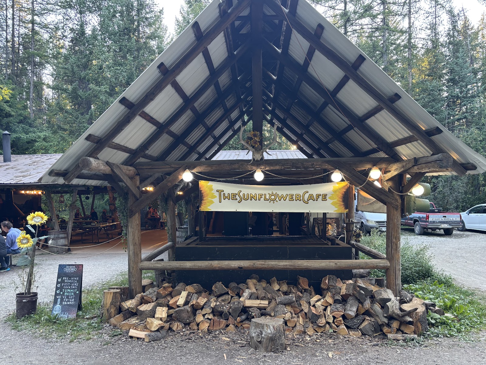 Rustic timber pavilion houses a seasonal cafe in Glacier National Park, with stacked firewood and sunflowers decor.