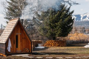 A-frame wooden sauna hut with a smoky chimney and stacked firewood in Glacier National Park, Montana.