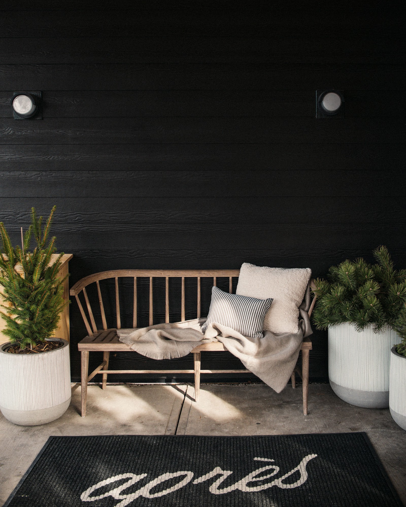 Apres Whitefish entrance at Glacier National Park features a wooden bench, striped cushion, soft throw, and potted evergreens against a dark wall.