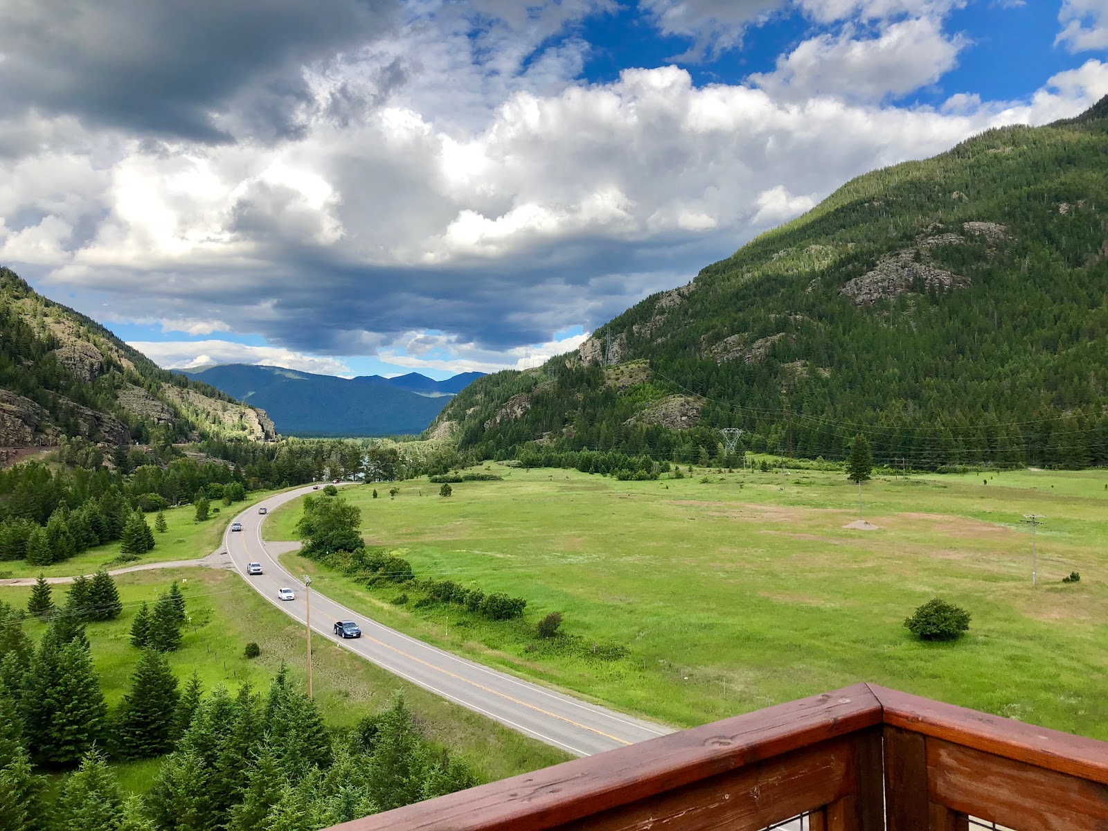 Glacier National Park valley view with winding road, green meadows, and forested mountains in the distance.