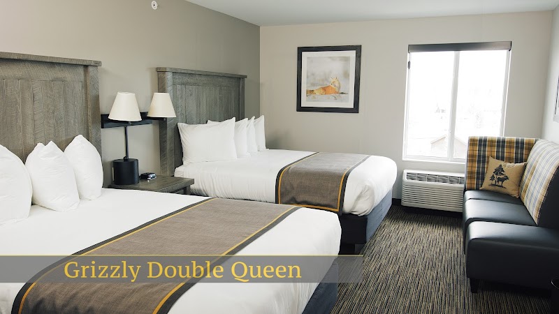 Two queen beds with white linens and brown runners, lamps, framed art, window view, plaid chair in Yellowstone National Park lodging.