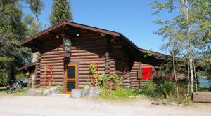 Log cabin office in Glacier National Park with a blue sign, orange-framed door, bikes to the left, and a lake visible beyond trees.
