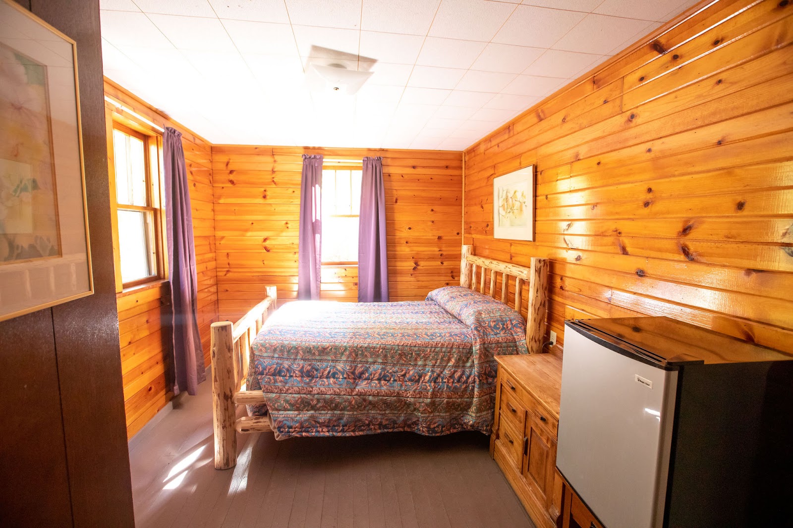 Cozy log cabin bedroom in Glacier National Park with a rustic wood bed, patterned quilt, purple curtains, and a small fridge.