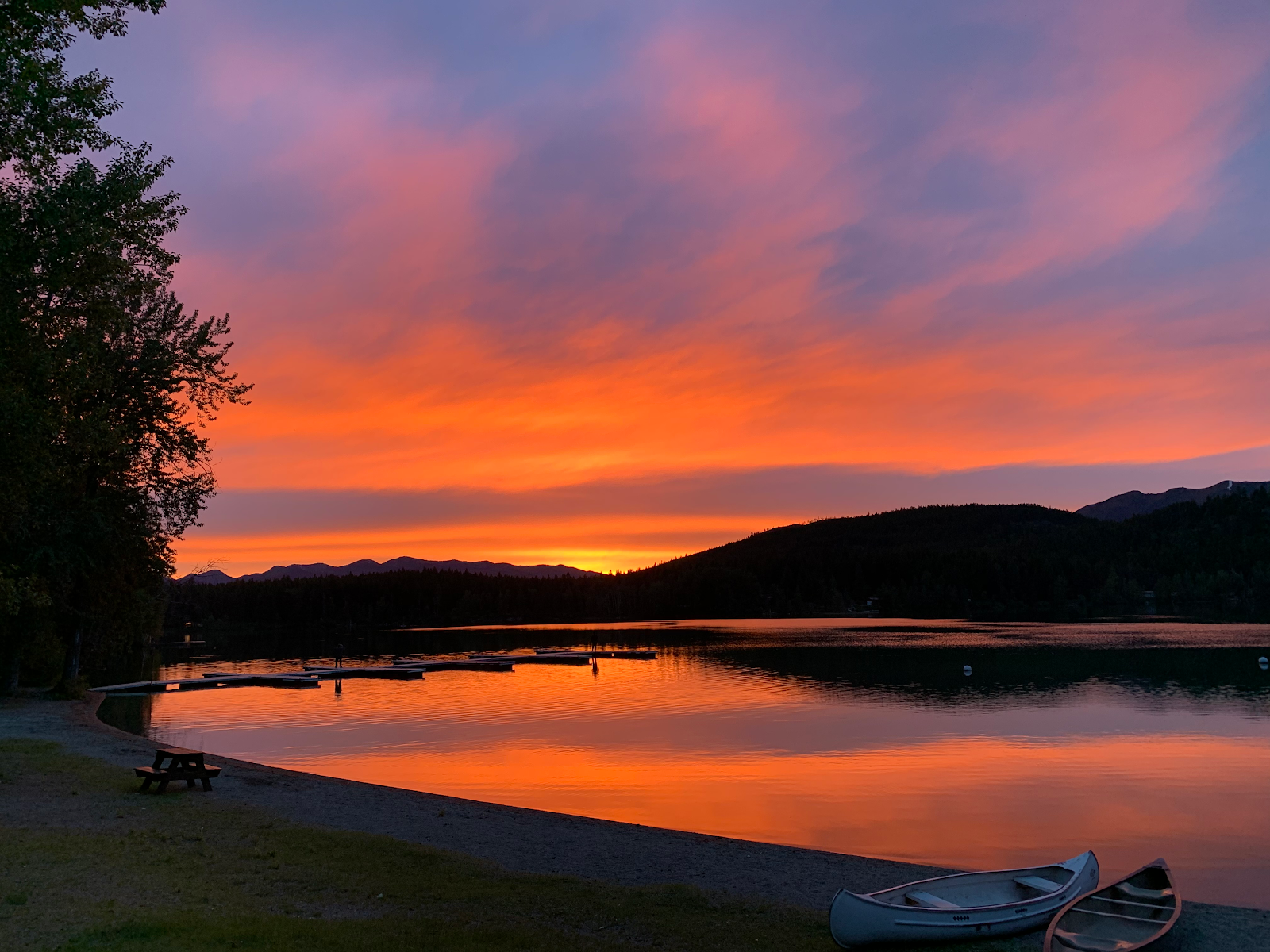 Sunset over a calm lake with orange-pink sky and mountain silhouettes; a dock, canoe, and pebbled shore with a picnic table in Glacier National Park.