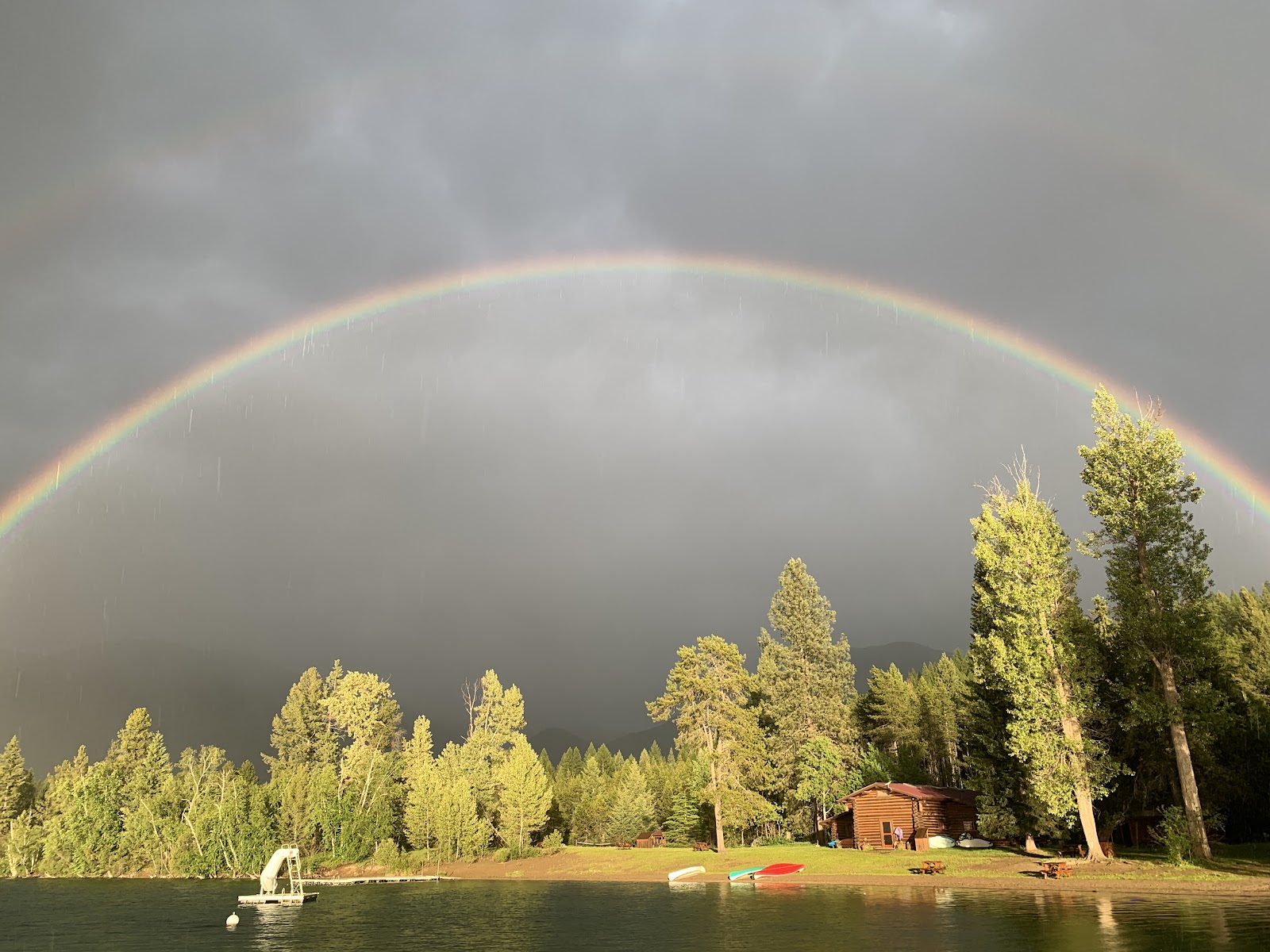 Calm lakefront at Glacier National Park with a wooden cabin, boats on the shore, and a double rainbow arching over tall pines.