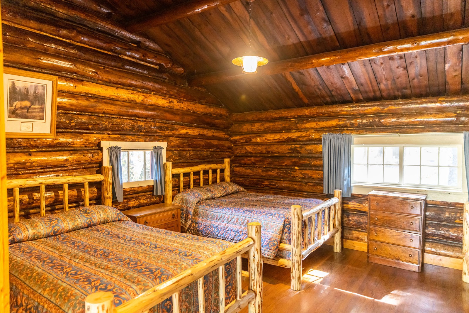 A rustic log cabin bedroom in Glacier National Park with two wooden twin beds, exposed log walls, a small window with blue curtains, and a wooden dresser.