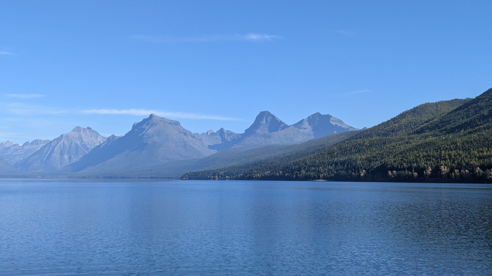 Station 8 at Glacier National Park shows a tranquil blue lake and forested slopes with rugged peaks under a clear sky.