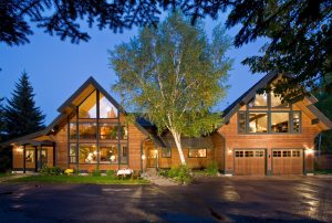 Glacier National Park lodge exterior glows warmly at dusk, with A-frame wings, wooden siding, and a central birch tree.