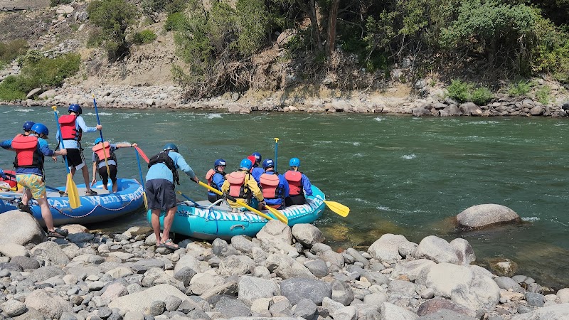 Rafting group in blue inflatable rafts with helmets and life jackets along a rocky Yellowstone National Park riverbank.