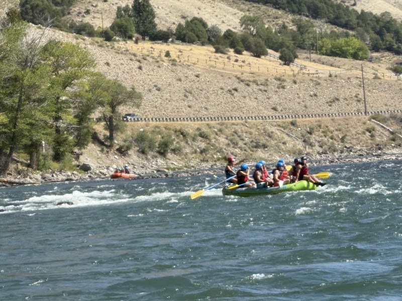 Yellowstone National Park: a bright green raft carries six paddlers in helmets through whitewater, with an orange raft visible ashore.