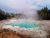 Turquoise hot spring pool with mineral crust, steam rising, encircled by pines in Norris Basin, Yellowstone National Park.