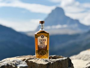 Logan Pass whiskey bottle sits on a rugged rock with Glacier National Park peaks in the distance.