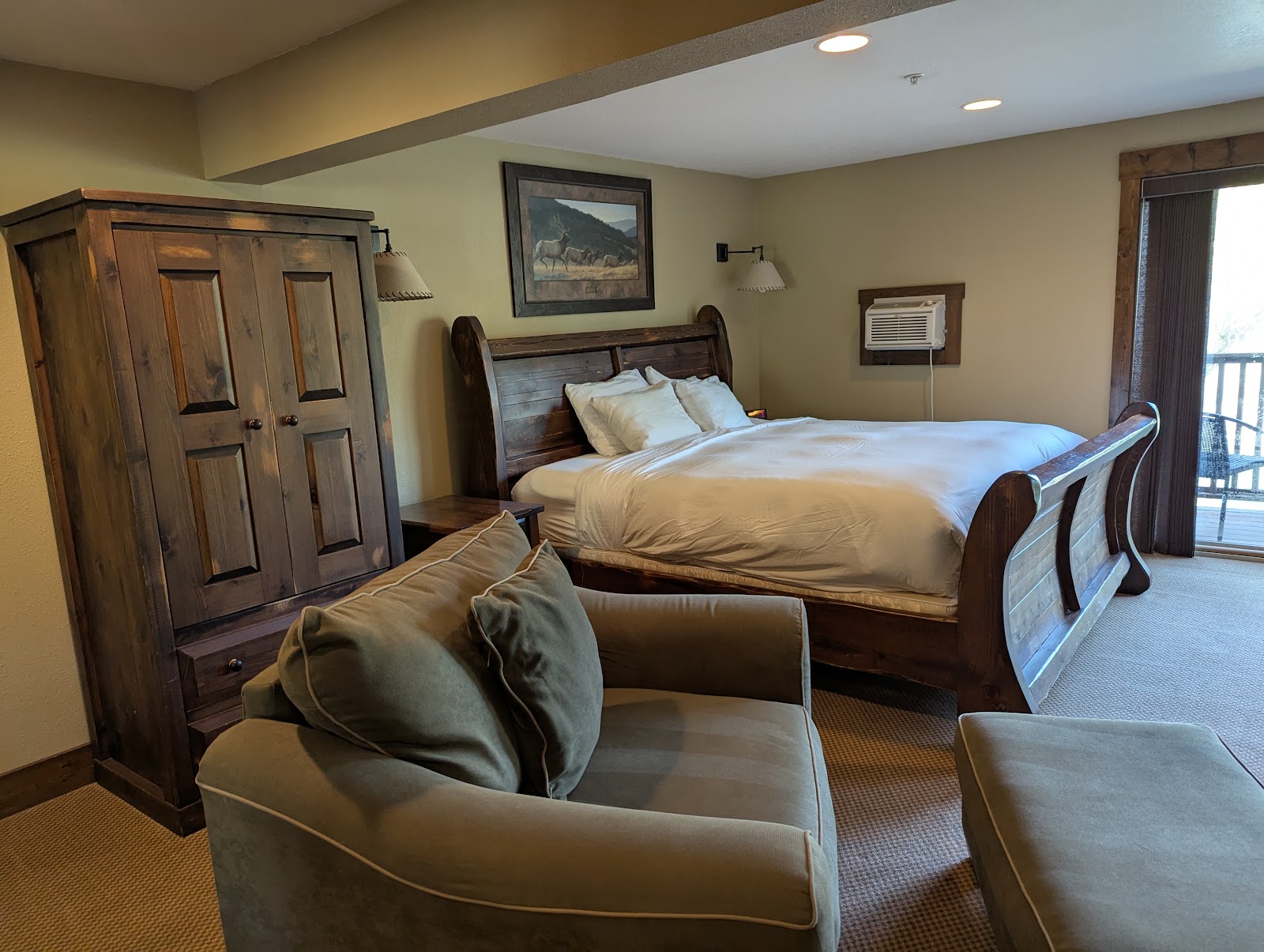 Cozy Glacier National Park lodge bedroom with a large carved wood bed, armoire, and a seating area by a private balcony.