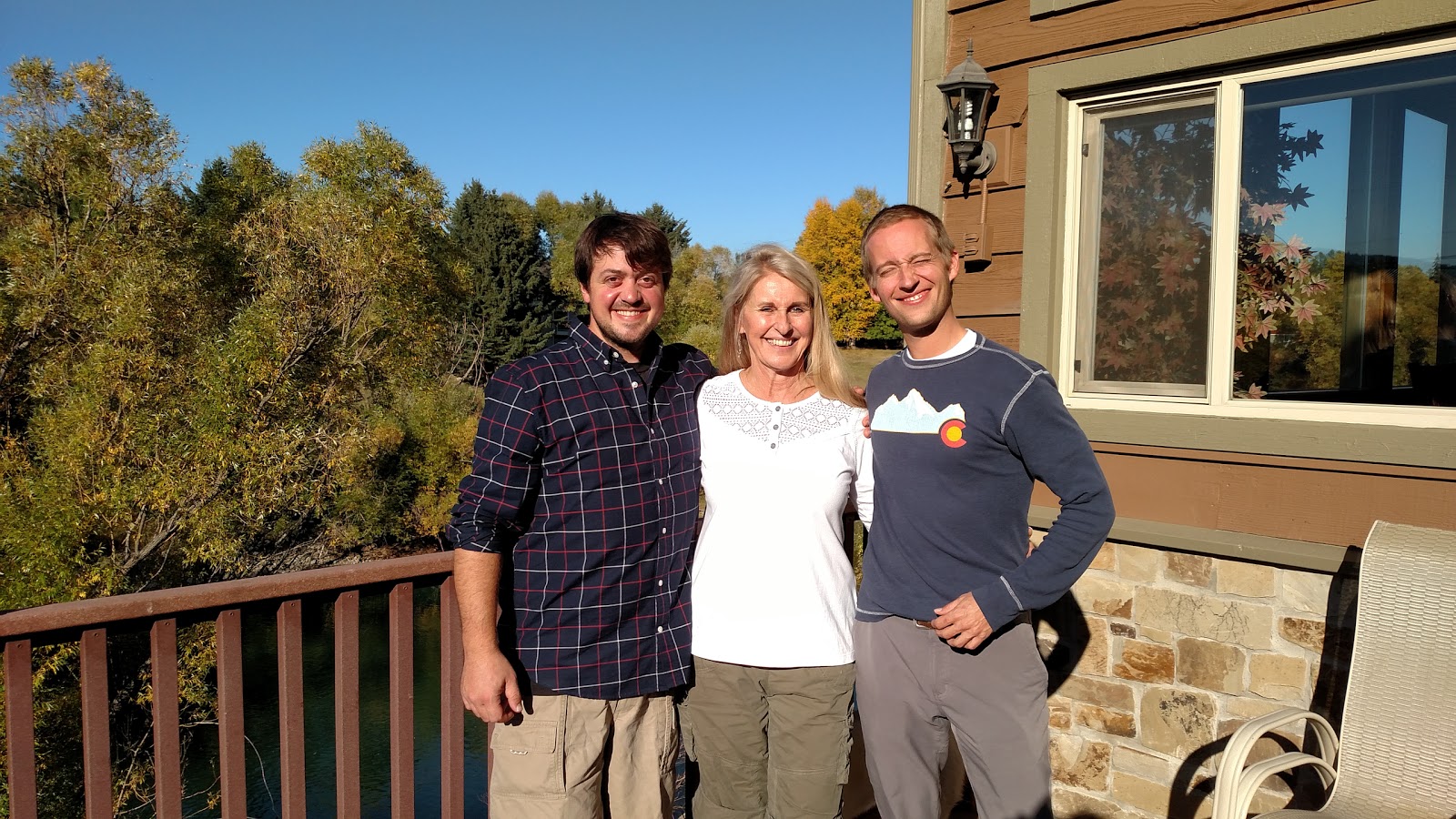 Three people stand on a lodge deck at Glacier National Park during fall foliage on a sunny afternoon.
