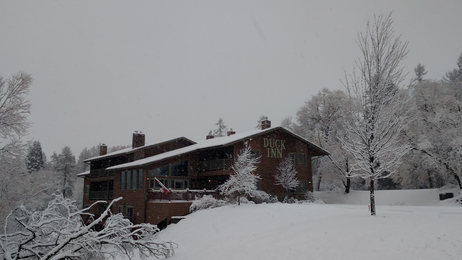 Snow covers a lodge exterior with a wraparound deck and surrounding bare, snow-laden trees in Glacier National Park.