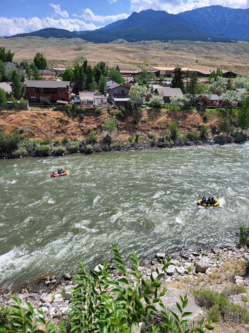 Riverside cottages line a rocky riverbank with a fast-moving green river, trees, and distant mountains in Yellowstone National Park.