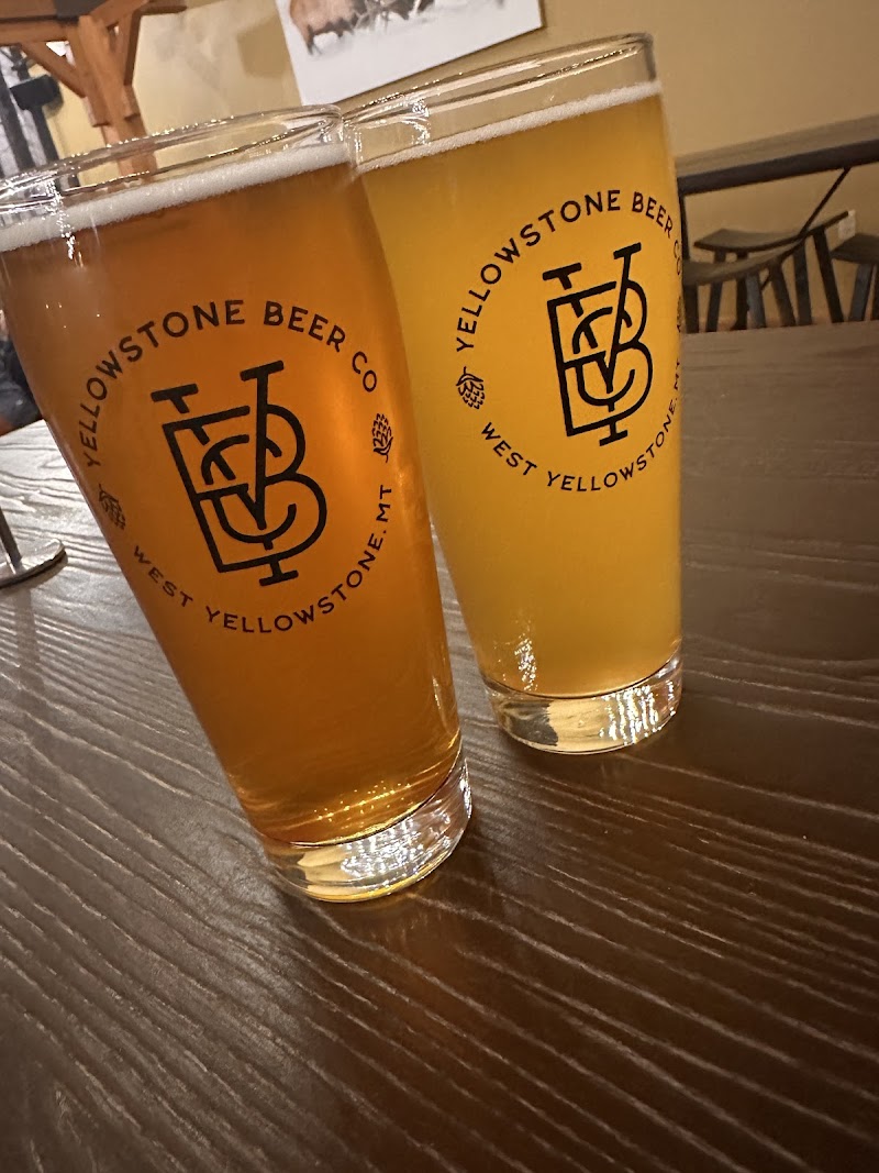 Two amber beers in tall glasses on a dark wood table with circular West Yellowstone MT logos, in Yellowstone National Park.