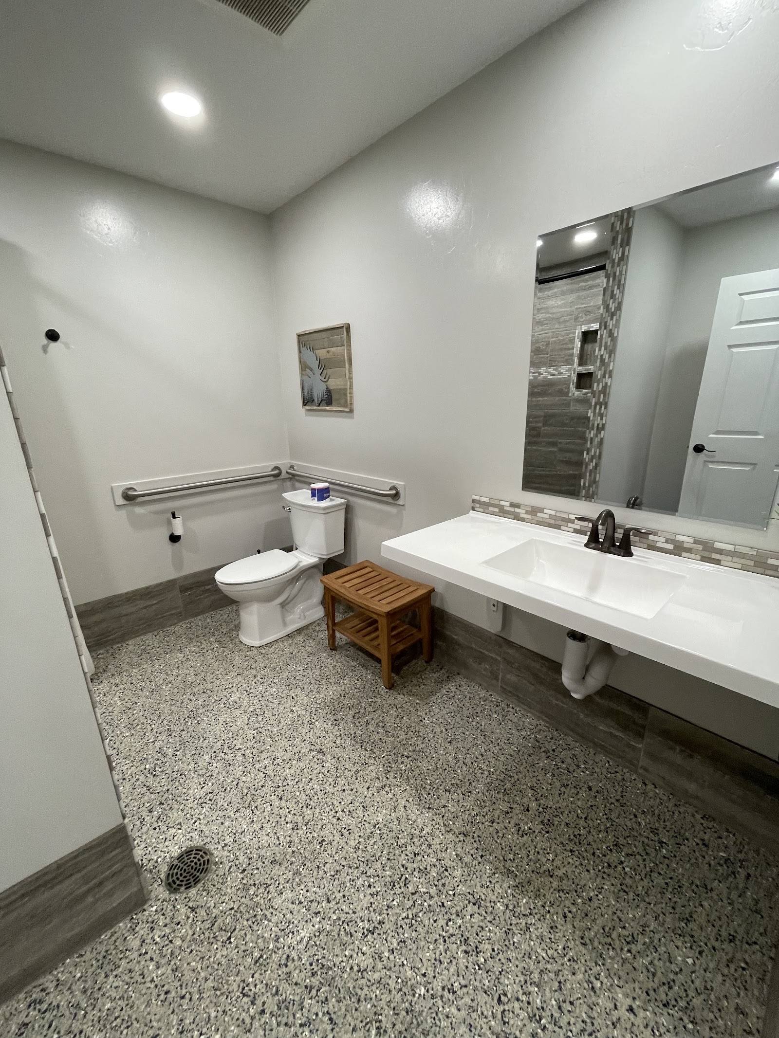 ADA-accessible campground bathroom with a long white sink, mirror, toilet, grab bars, wooden stool, and speckled floors in Glacier National Park.