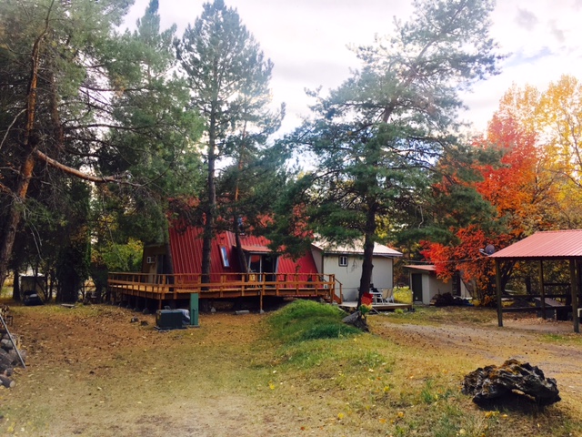 Autumn campground in Glacier National Park with red cabins, a wooden deck, tall pines, and colorful fall foliage.
