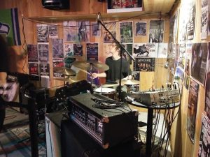 Interior of a bar-like music venue in Glacier National Park with a drummer on stage and posters lining the wood-paneled walls.