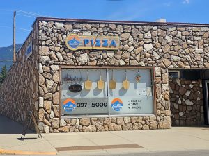 Exterior of a rustic stone building housing a pizza restaurant in Glacier National Park, with blue sky overhead.