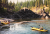 Kayakers paddle near a rocky shoreline along a glacier-fed river in Glacier National Park, surrounded by forested walls.