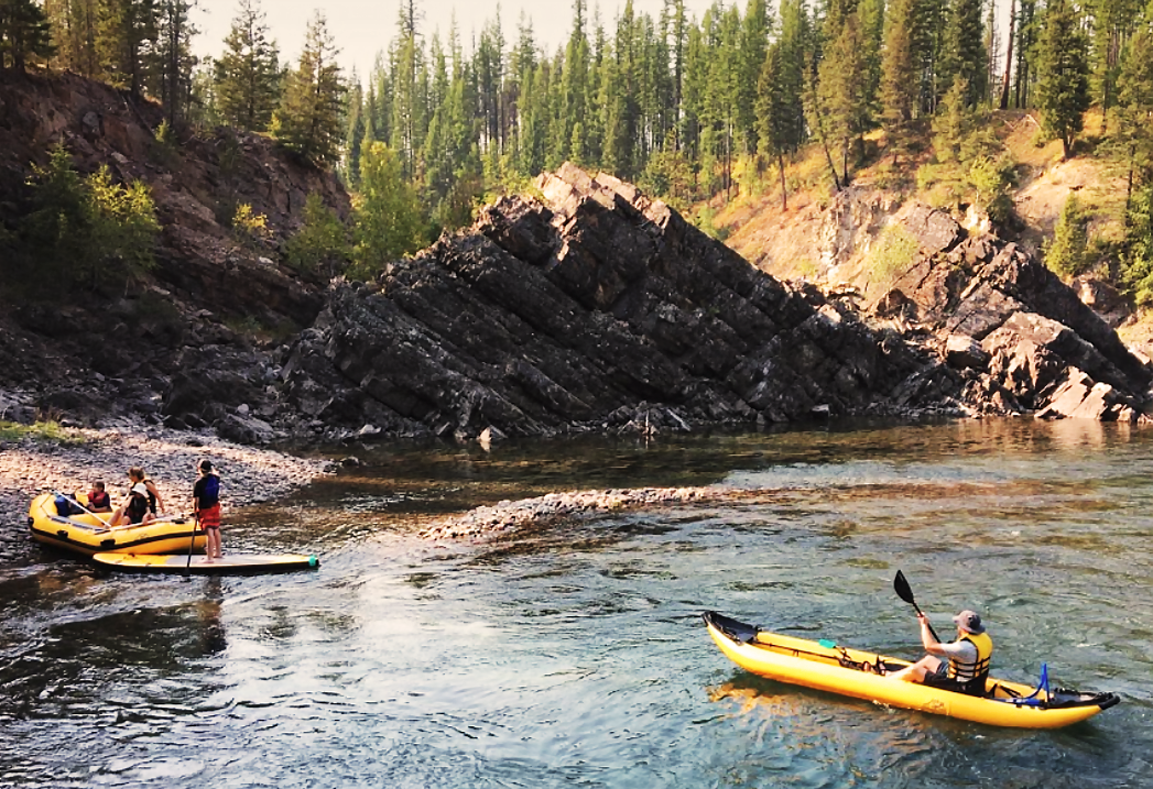 Kayakers paddle near a rocky shoreline along a glacier-fed river in Glacier National Park, surrounded by forested walls.