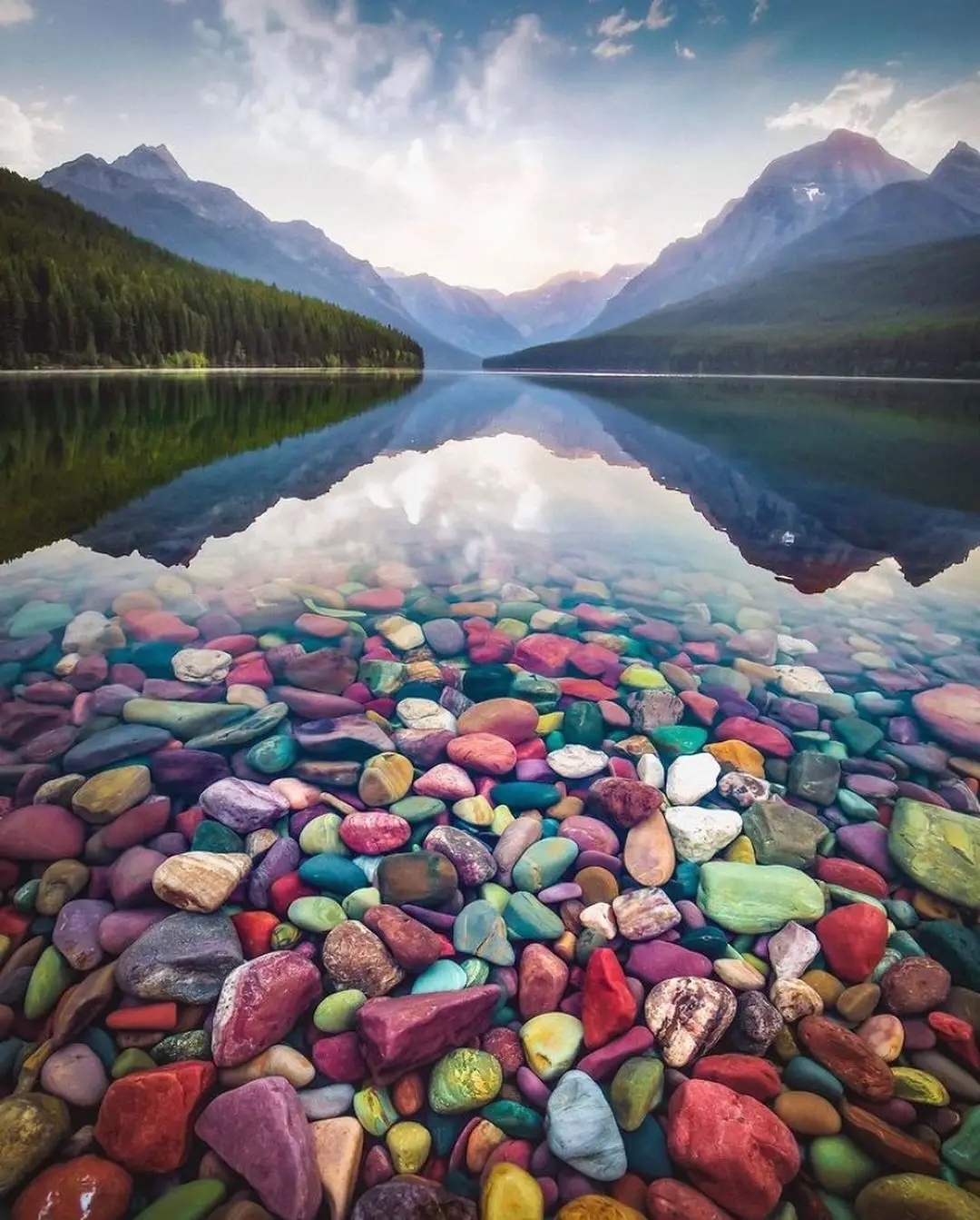 Colorful smooth argillite stones — red, green, orange, and purple — fill the foreground at Lake McDonald with Glacier National Park mountains reflected in the background