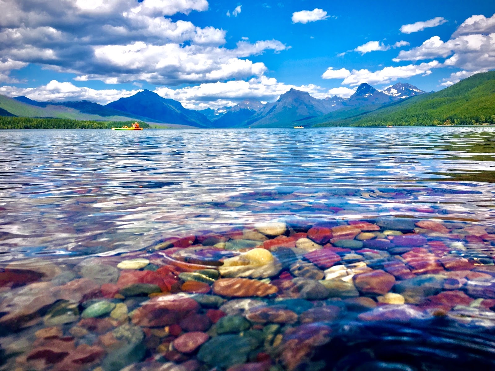 Clear Lake McDonald water gently lapping colorful argillite pebbles at the shoreline with distant snow-capped peaks in Glacier National Park