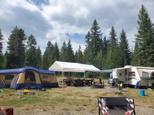Campground scene with a blue and tan tent, white canopy over picnic tables, an RV, folding chairs, and tall pines at Glacier National Park.