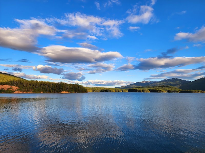 Calm blue lake reflects a forested shore and distant mountains under a bright sky in Glacier National Park.