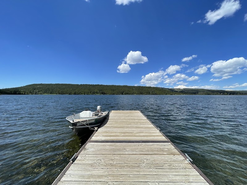 Wooden dock extends into a lake with a small motorboat moored at the edge, pine forest and sky in Yellowstone National Park.