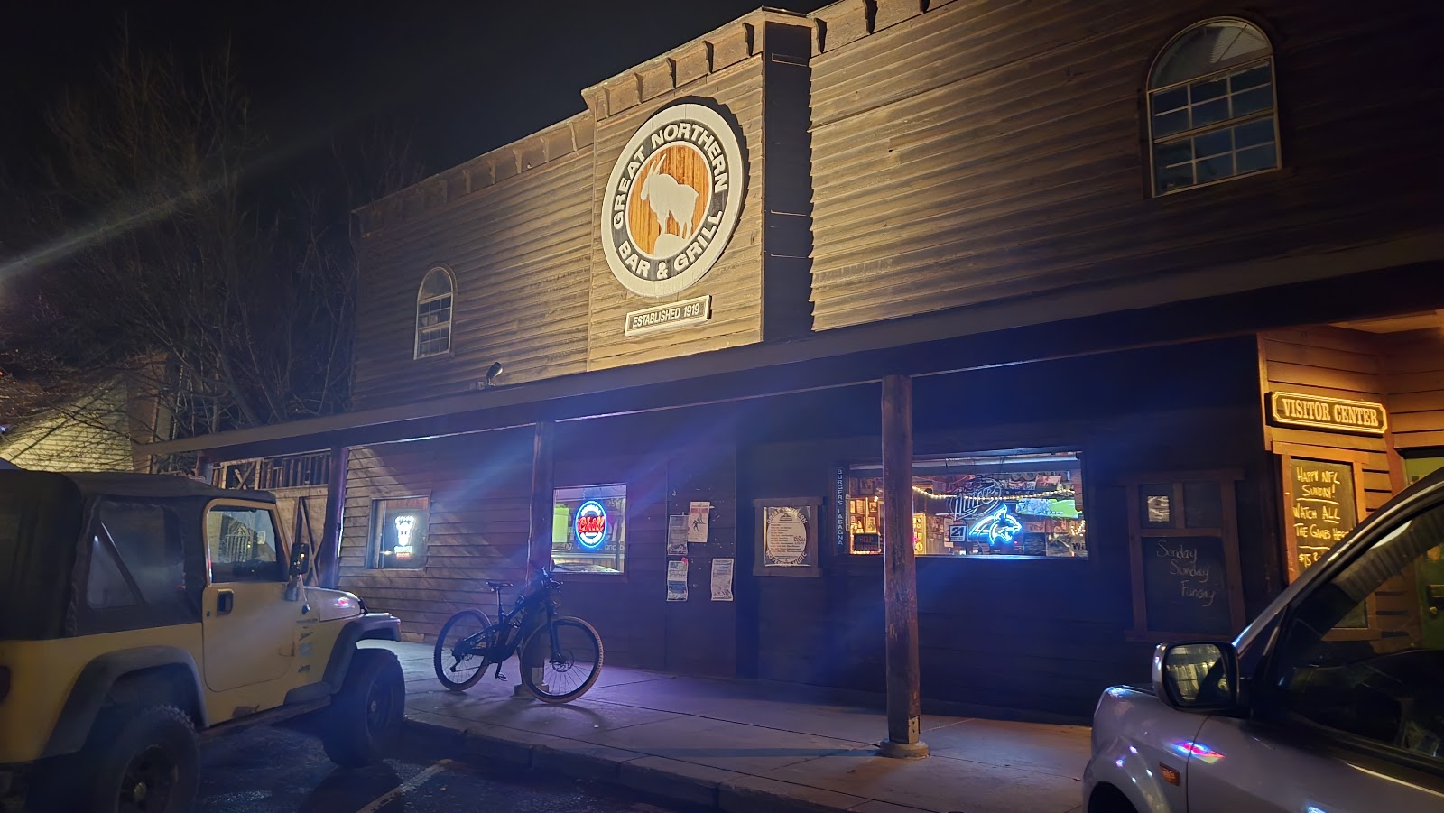 Exterior view of a rustic bar and grill in Glacier National Park, lit at night with a large circular sign and wooden facade.