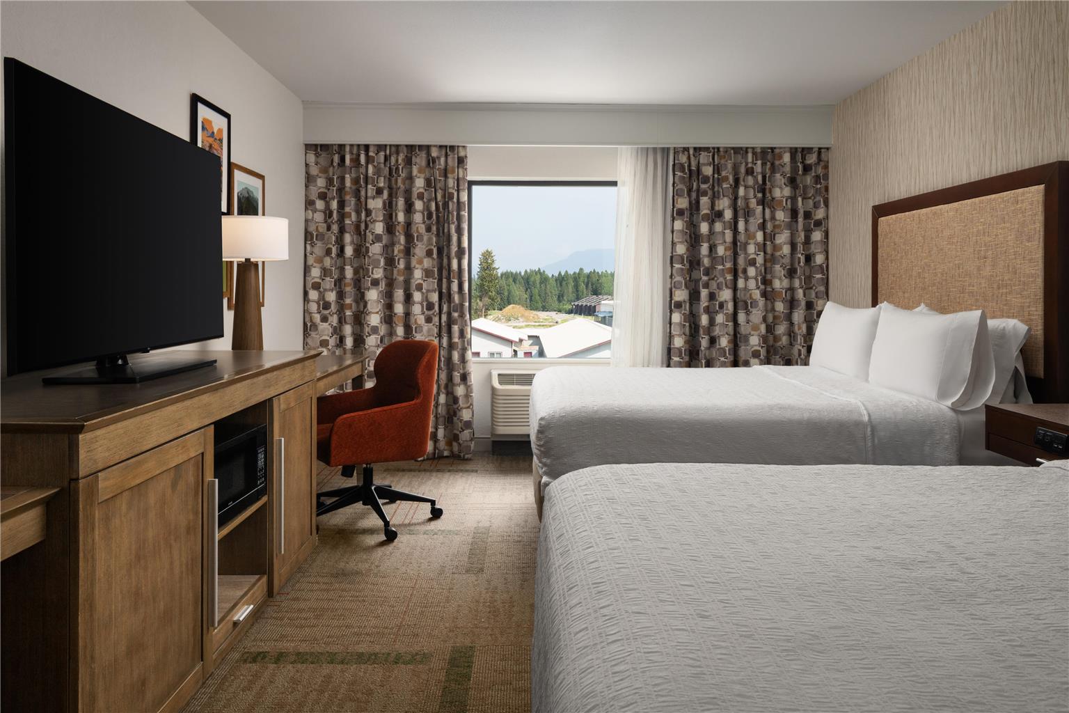 Bright hotel room with two queen beds and a window view of the town and distant mountains near Glacier National Park.