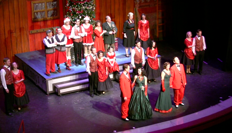 Actors in red and green period costumes perform on a wooden stage with a Christmas tree, Yellowstone National Park.