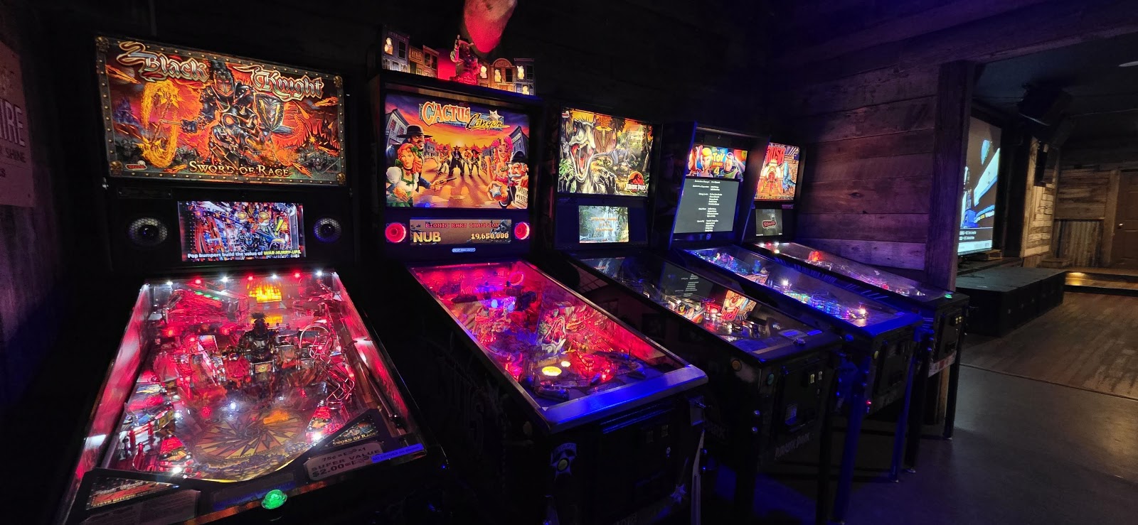 Inside a dimly lit arcade at Glacier National Park, neon-lit pinball machines line a wooden-walled room.