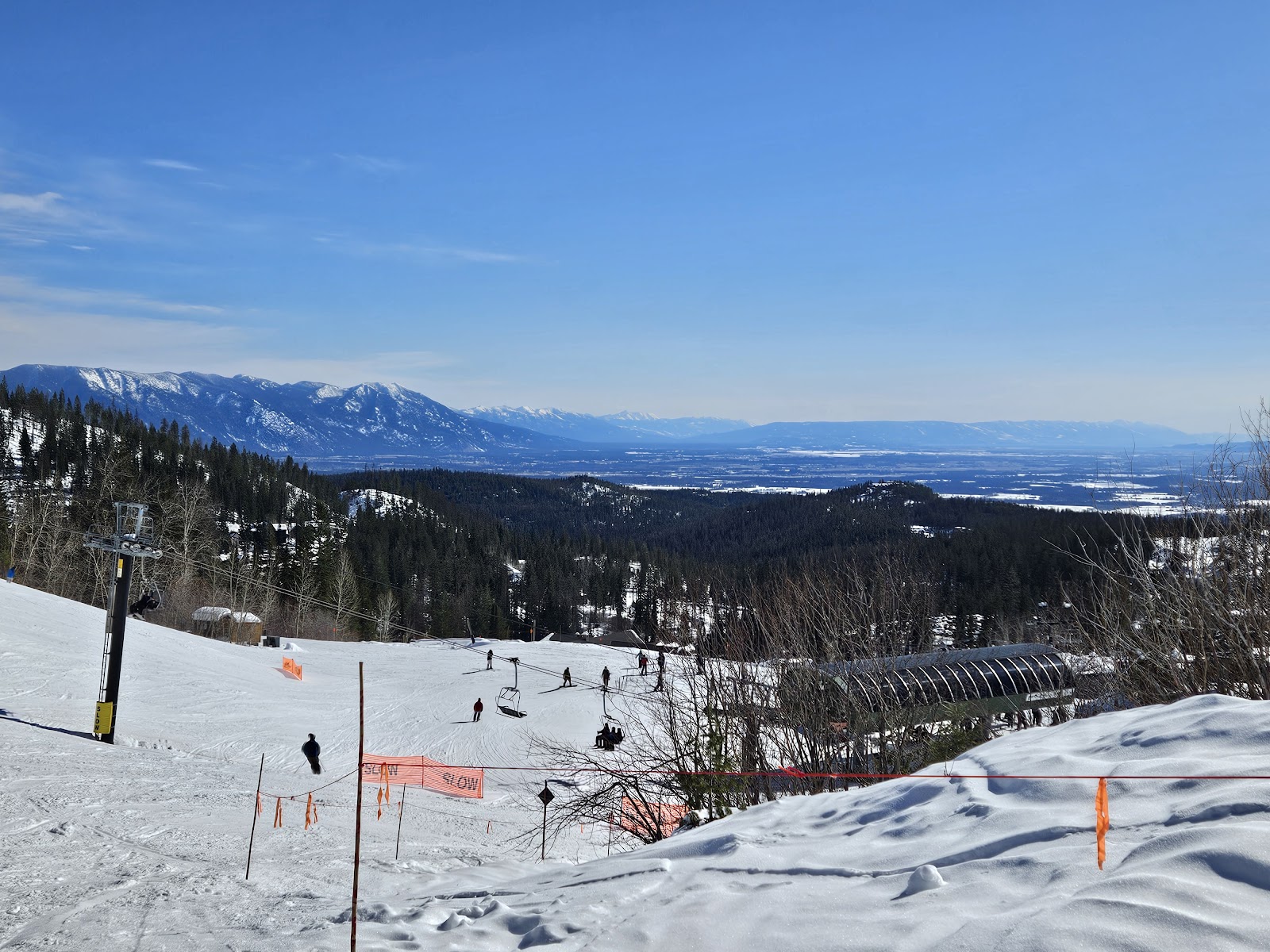 Snowy ski slope with a chairlift and pine forest below Glacier National Park, distant mountains under a clear blue sky.