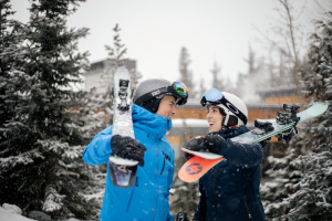 Two skiers in bright jackets chat in a snowy Glacier National Park area near Whitefish Mountain Resort, with gear strapped to their shoulders.