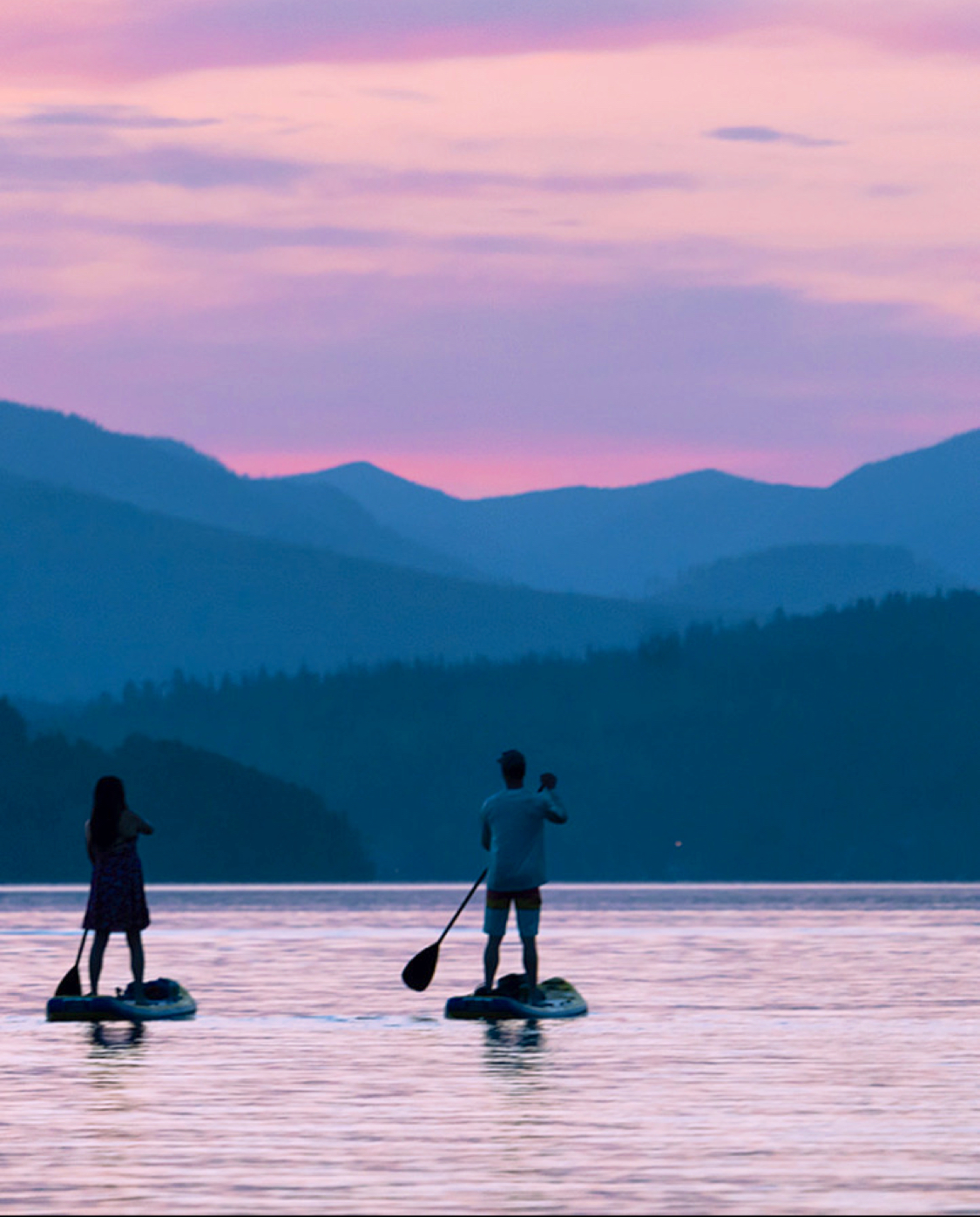 Paddleboarders glide on a calm lake at sunset with blue mountain silhouettes behind, in Glacier National Park.