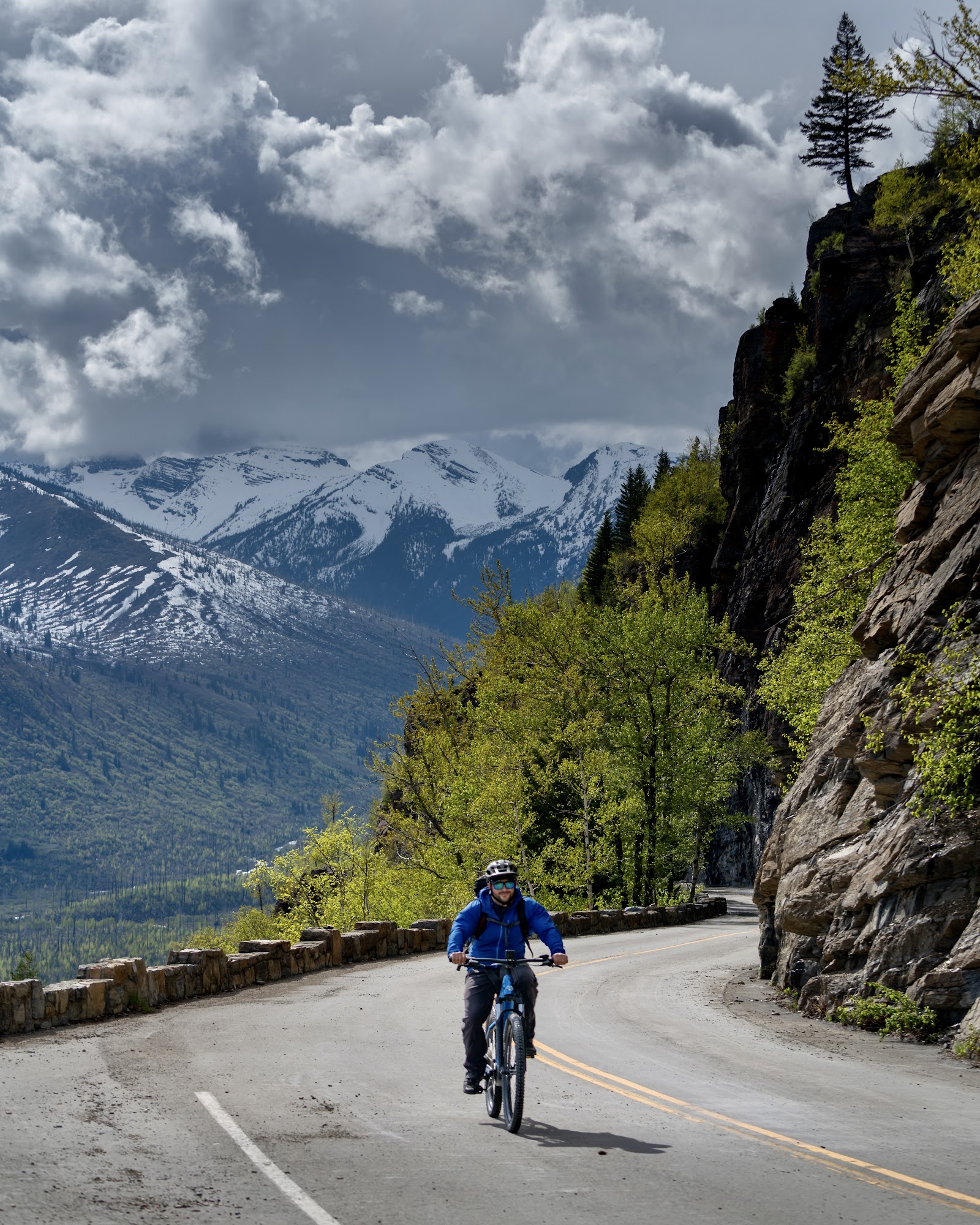 Biker pedals along Going-to-the-Sun Road, Glacier National Park, with snow-dusted peaks and dramatic cliffside scenery.