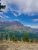 Aster Park Overlook view in Glacier National Park showing rugged mountain slopes, green forests, and a dark lake below under a blue sky.