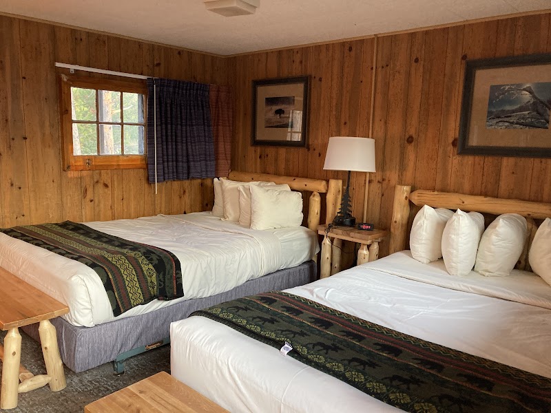 Rustic, wood-paneled lodge room in Yellowstone National Park with two queen beds, white linens, and a bedside lamp.